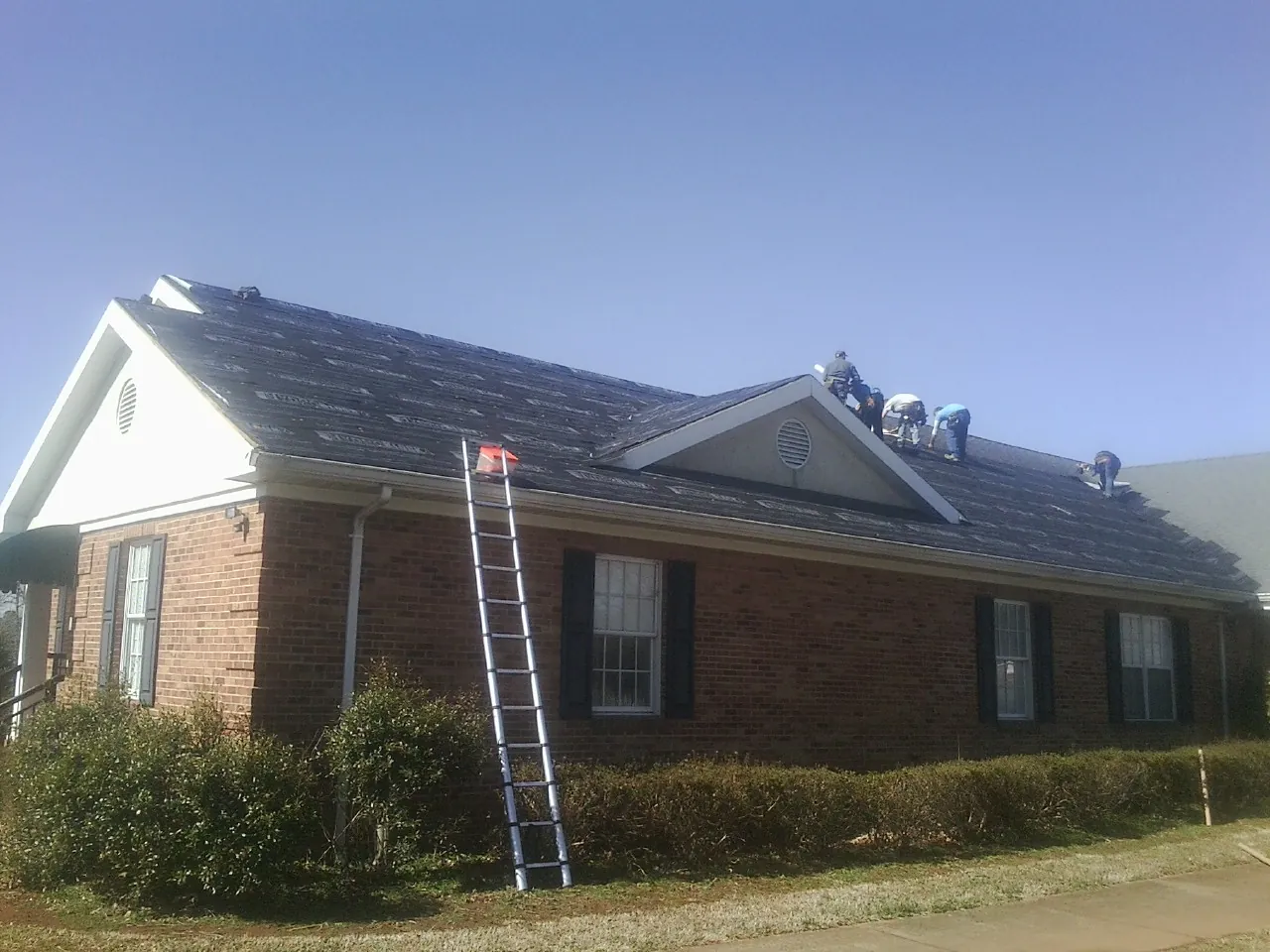 Crew tearing shingles off a brick ranch roof with a ladder set — Emergency Roof Repair work by our crew in Port Sanilac