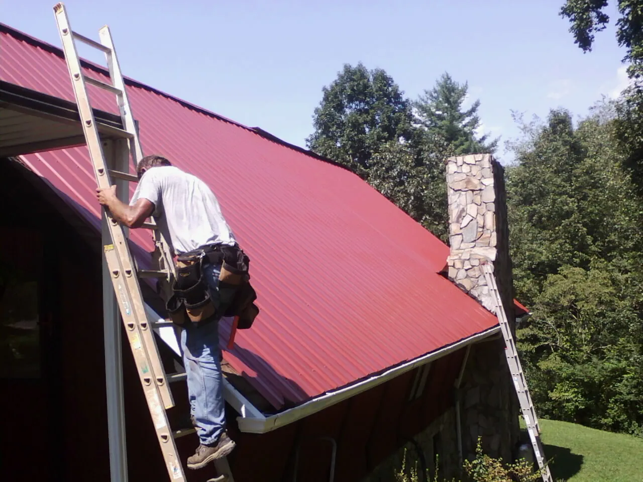 Roofer on a ladder beside a red metal roof and stone chimney — Soffit & Fascia Repair by Roofing Brothers in Port Sanilac