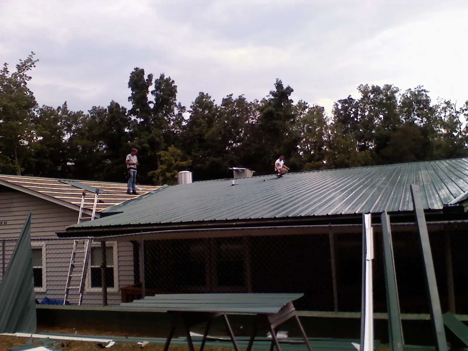 Two roofers installing green metal panels above a covered porch in Port Sanilac